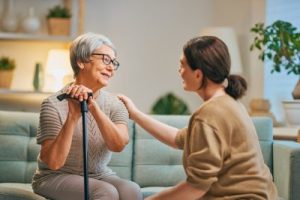 Female caregiver crouches to comfort seated female senior with a cane