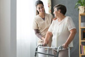 Female caregiver assists female senior while walking with walker